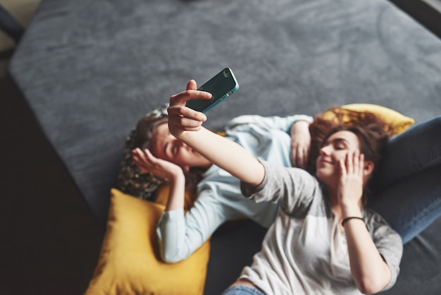 Duas irmãs gêmeas sorridente fofo segurando o smartphone e fazendo selfie. meninas mentem no sofá posando e alegria