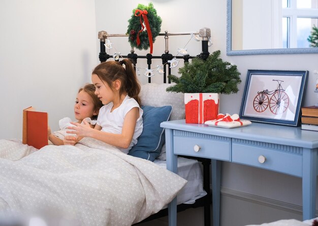 Foto duas irmãs pequenas estão lendo um livro na cama enquanto estão em um quarto decorado com decorações de natal e guirlandas