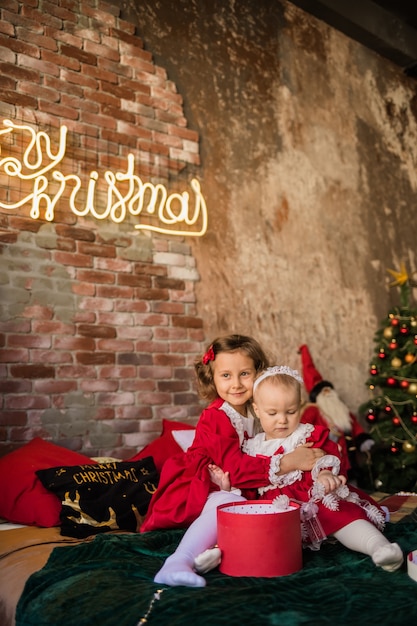 Duas irmãzinhas em vestidos de festa vermelhos sentam-se, abraçam-se na cama e brincam com as caixas da árvore de Natal