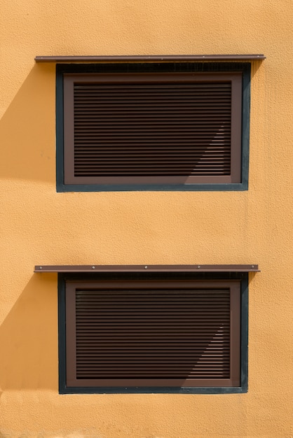 Duas janelas marrons com persianas fechadas na parede amarela. Imagem externa do edifício industrial