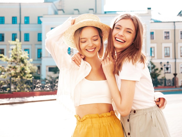Duas jovem bela mulher hipster sorridente em roupas da moda de verãoMulheres despreocupadas sexy posando no fundo da rua no chapéu Modelos puros positivos se divertindo ao pôr do sol abraçando