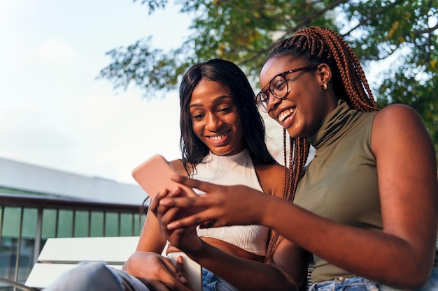 Duas jovens africanas se divertindo sentadas em um banco de parque com um conceito de celular da juventude