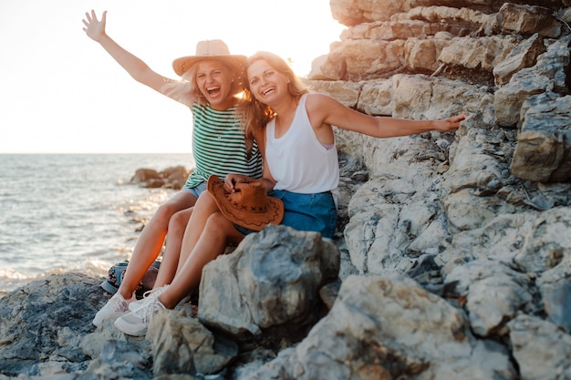 Duas jovens alegres em chapéus descolados na rocha na costa do mar. Paisagem do verão com menina, mar, ilhas e luz solar laranja.