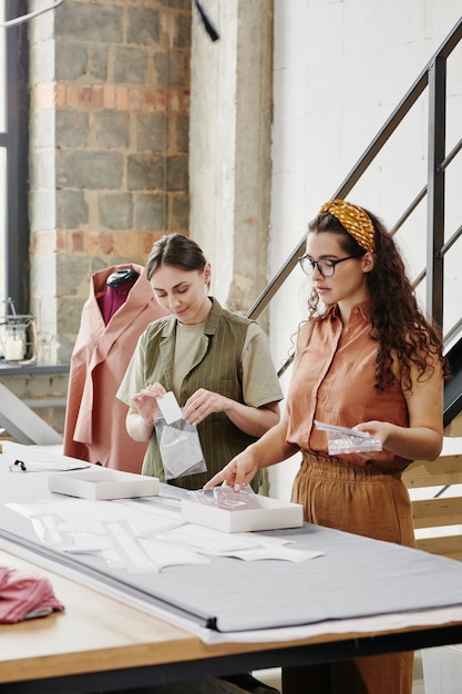 Duas jovens alfaiates em roupas casuais carregando ombreiras novas para casacos, jaquetas e vestidos, enquanto estavam de pé junto à mesa no estúdio