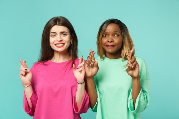 Foto duas jovens amigas afro-americanas europeias em roupas verdes rosa, mantendo os dedos cruzados isolados no retrato de estúdio de fundo de parede azul turquesa. conceito de estilo de vida de pessoas. espaço de cópia de mock up