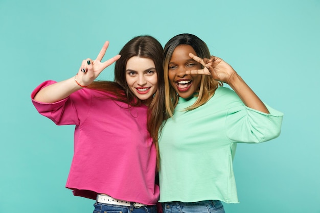 Duas jovens amigas europeias, afro-americanas em roupas verdes rosa, mostrando sinal de vitória isolado no fundo da parede azul turquesa, retrato de estúdio. conceito de estilo de vida de pessoas. mock up espaço de cópia.