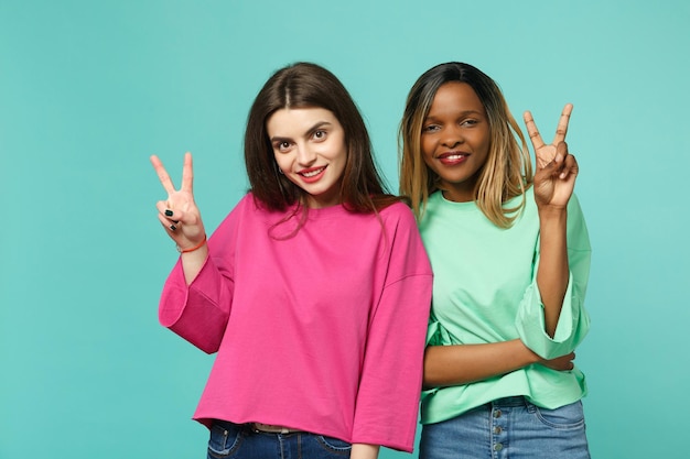 Foto duas jovens amigas europeias, afro-americanas em roupas verdes rosa, mostrando sinal de vitória isolado no fundo da parede azul turquesa, retrato de estúdio. conceito de estilo de vida de pessoas. mock up espaço de cópia.