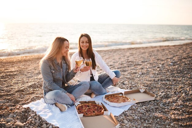 Duas jovens amigas lindas se divertindo com pizza e vinho na praia ao longo da costa do mar ao ar livre na luz do sol. Temporada de férias de verão. Amizade. Felicidade. As irmãs passam um tempo juntas na costa.