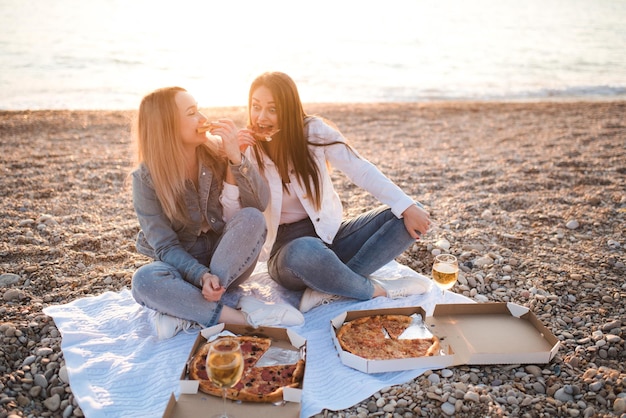 Duas jovens amigas lindas se divertindo com pizza e vinho na praia ao longo da costa do mar ao ar livre na luz do sol. Temporada de férias de verão. Amizade. Felicidade. As irmãs passam um tempo juntas na costa.