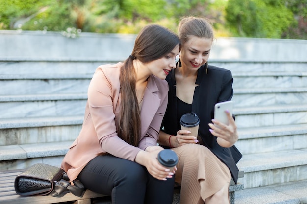 Duas jovens amigas sorridentes em roupas de negócios estão olhando para a tela do smartphone enquanto estão sentadas em um banco no parque