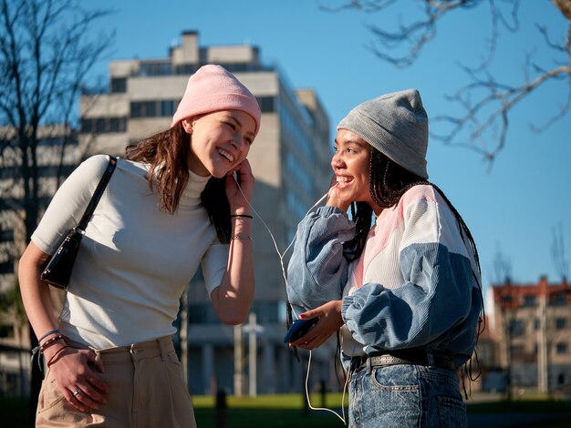 Duas jovens amigas vestindo roupas casuais e usando um smartphone com fones de ouvido