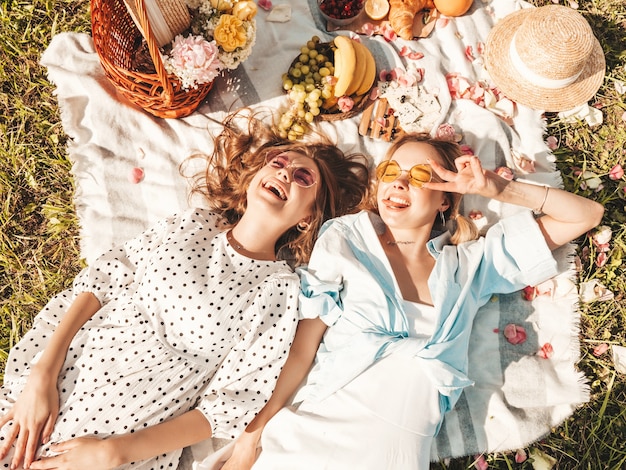 Duas jovens belas garotas hippie sorridentes em vestido de verão da moda e chapéus. Mulheres despreocupadas fazendo piquenique do lado de fora.