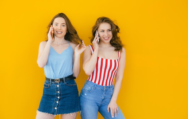 Duas jovens bonitas surpresas usando telefone celular juntas sobre fundo amarelo