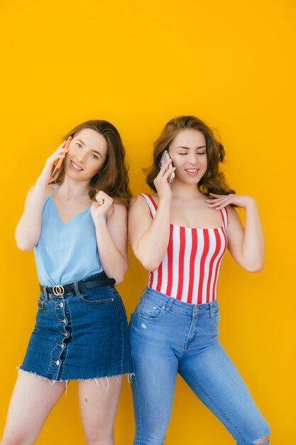 Duas jovens bonitas surpresas usando telefone celular juntas sobre fundo amarelo