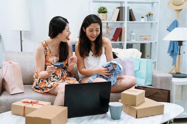 duas jovens chinesas asiáticas desempacotando encomendas em casa sentadas no sofá ao redor de caixas e sacolas de compras. amigas de meninas com cartão de crédito e computador portátil comprando compra na venda pela internet