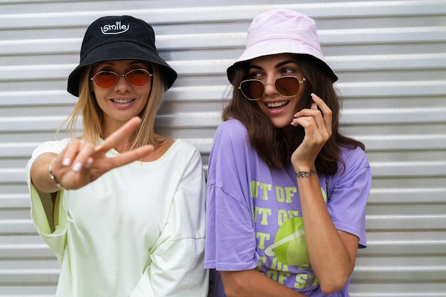 Duas jovens e elegantes hipster sorridentes amigas no panamá da moda e óculos de sol da moda posando ao ar livre se divertindo rindo