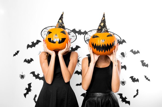 Duas jovens em vestidos pretos e chapéus de bruxa seguram abóboras de Halloween em seus rostos.