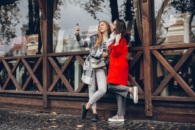 Duas jovens empresárias elegantes em pé em uma cidade de outono e usam o telefone