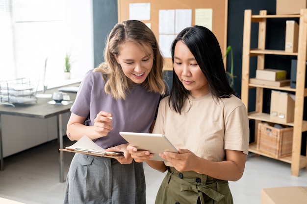 Duas jovens empresárias felizes com um tablet digital examinando pedidos on-line enquanto uma delas compara os dados com os do papel