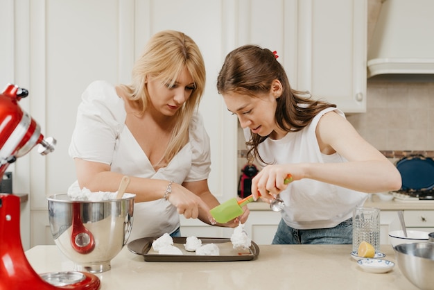 Duas jovens estão colocando diligentemente o merengue recém-batido em uma bandeja com uma colher e uma escápula na cozinha. as meninas estão se preparando para cozinhar uma deliciosa torta de merengue de limão.