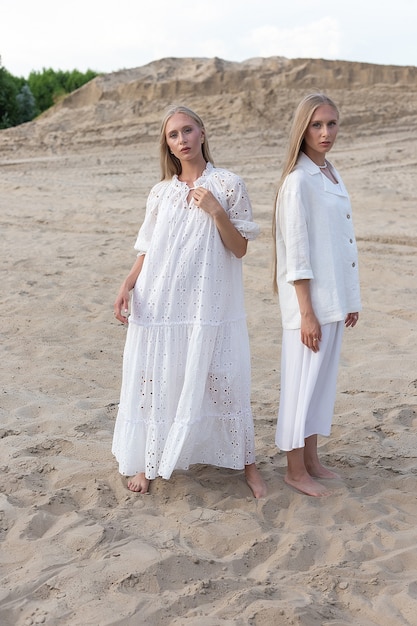 Duas jovens gêmeas atraentes com cabelo comprido passando um tempo juntos ao ar livre no verão, posando em uma pedreira de areia em um vestido branco, saia, jaqueta