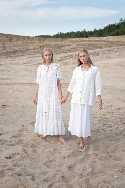 Duas jovens gêmeas bonitas com longos cabelos loiros posando em uma pedreira de areia em elegantes vestidos brancos