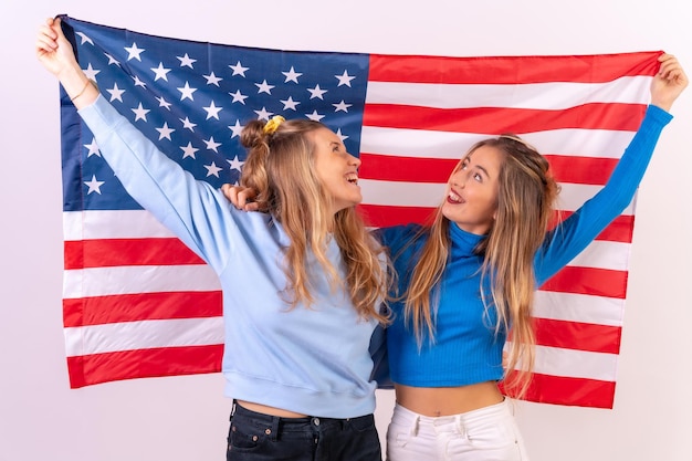 Duas jovens loiras caucasianas com bandeira americana sorrindo enquanto seguram a bandeira isolada em um fundo branco