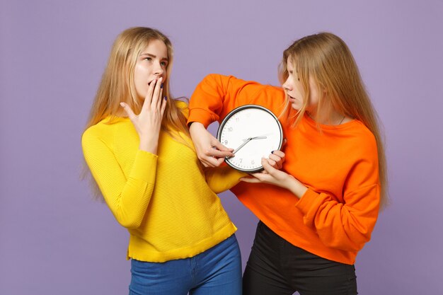 Duas jovens loiras gêmeas irmãs em roupas coloridas vivas, segurando o relógio redondo, movendo as setas isoladas na parede azul violeta pastel. conceito de estilo de vida familiar de pessoas.