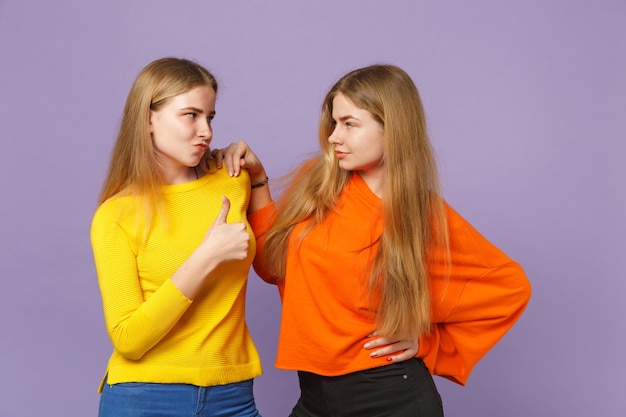 Duas jovens loiras gêmeas irmãs garotas em roupas coloridas vivas, olhando uma para a outra, aparecendo o polegar isolado na parede de azul violeta pastel. Conceito de estilo de vida familiar de pessoas.