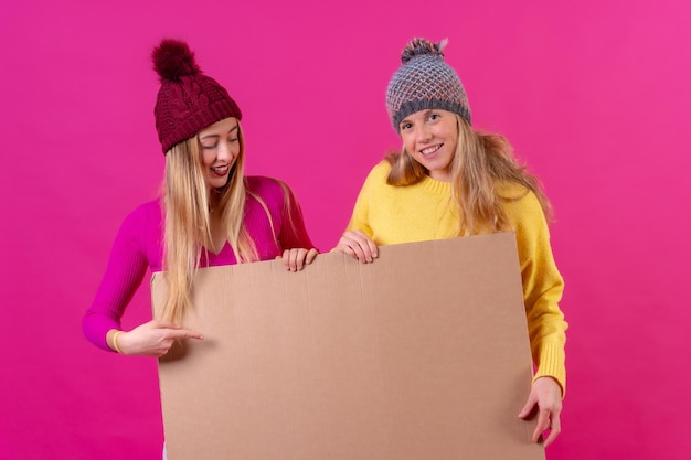 Duas jovens loiras segurando em suas mãos uma placa de venda isolada sobre um fundo rosa