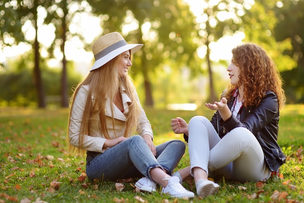 Duas jovens mulheres bonitas que falam ao sentar-se na terra no parque ensolarado.