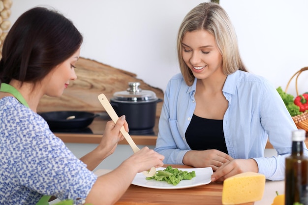 Duas jovens mulheres felizes estão cozinhando na cozinha amigos estão se divertindo enquanto preparam uma refeição saudável e saborosa