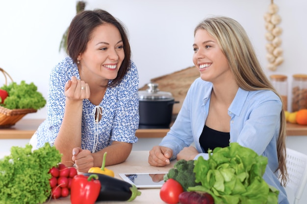 Duas jovens mulheres felizes estão fazendo compras on-line por computador tablet e cartão de crédito Amigos vão cozinhar na cozinha