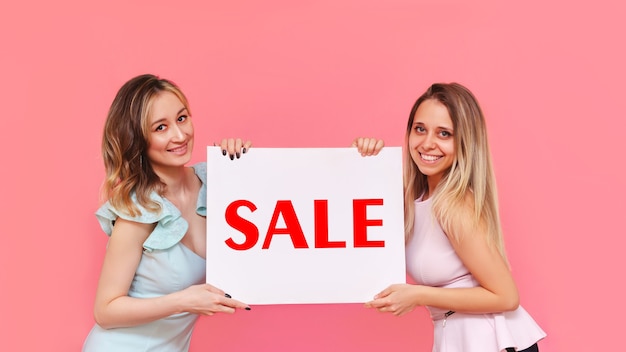 Foto duas jovens mulheres muito sorridentes segurando pôster branco com a palavra venda em um fundo rosa