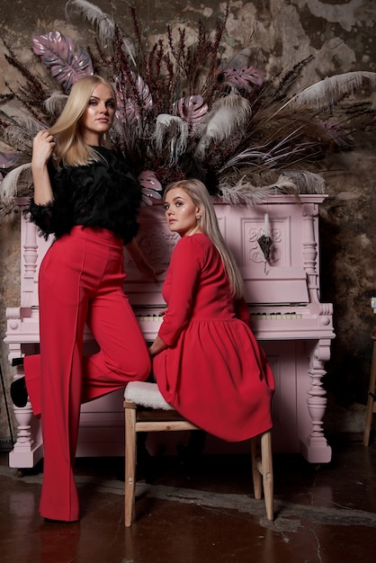 duas jovens mulheres sexy, com longos cabelos loiros elegantes e maquiagem glamourosa elegante e vestidos vermelhos perto do piano