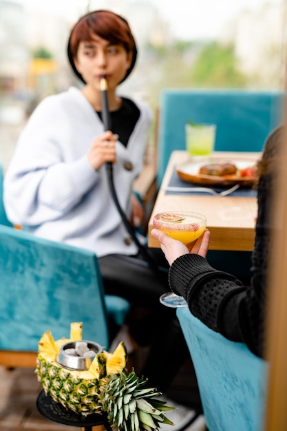 Duas jovens mulheres vestidas casuais bebendo coquetéis e fumando cachimbo de água no terraço de verão descansando