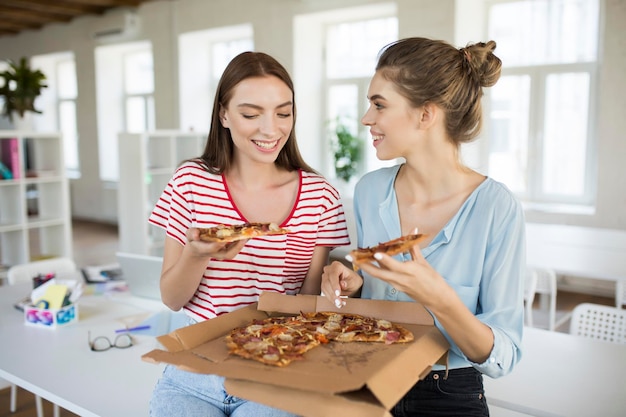 Duas jovens sorridentes sentadas na mesa conversando e comendo pizza alegremente enquanto passam tempo juntos no escritório moderno