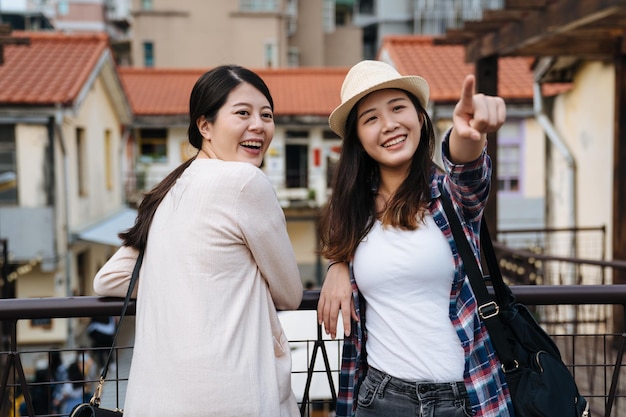 duas jovens turistas viajam no verão na pequena cidade do japão. mulher apontar para o céu com beauitful pôr do sol, mostrando o amigo encostado no corrimão, vire-se na pequena aldeia. sorrindo senhoras atraentes.