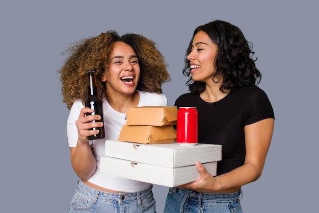 Duas jovens, uma latina e outra com cabelo afro, riem enquanto seguram pizzas e hambúrgueres