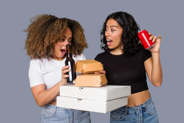 Duas jovens, uma latina e outra com cabelo afro, riem enquanto seguram pizzas e hambúrgueres