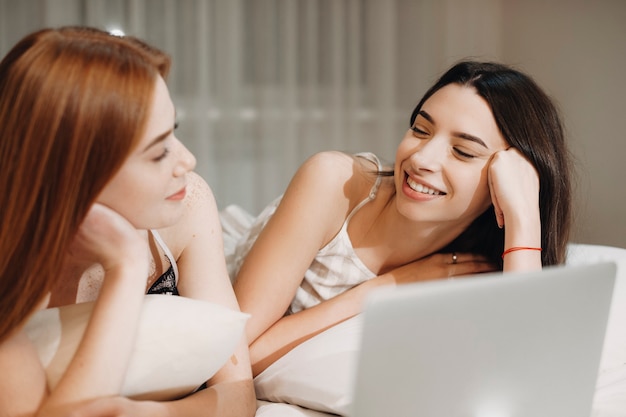 Duas linda jovem namorada se divertindo enquanto está encostado em uma cama com um laptop na frente do rosto, olhando um para o outro rindo.