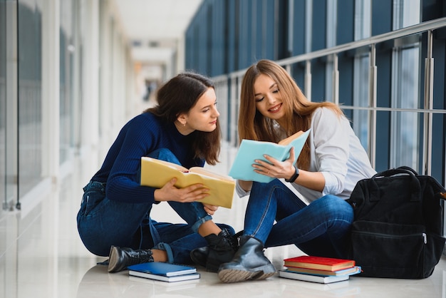 Duas lindas alunas com livros sentadas no chão no corredor da universidade