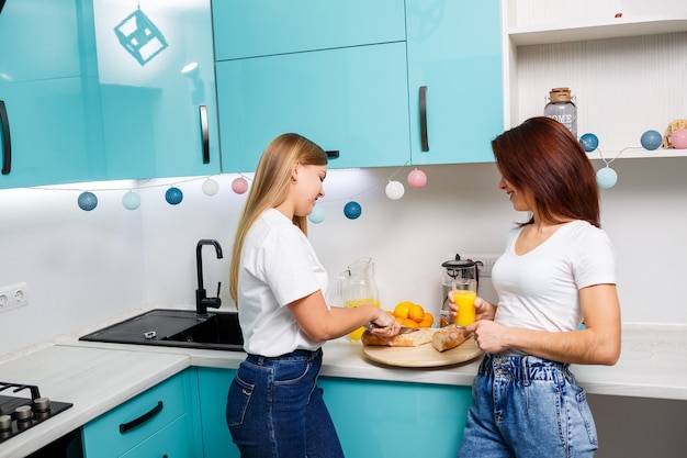 Duas lindas amigas jovens sentam-se à mesa da cozinha em casa, comem pão e bebem suco de laranja