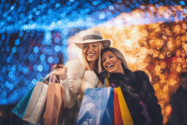 Foto duas lindas amigas sorridentes com muitos sacos coloridos estão se divertindo na hora das compras no feriado de natal no centro da cidade.