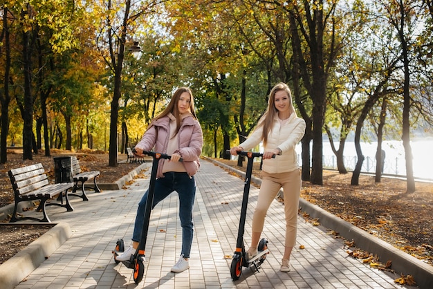 Duas lindas garotas andando de scooters elétricas no parque em um dia quente de outono