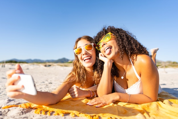 Foto duas lindas garotas deitadas na praia no verão se divertindo fazendo caretas com a língua de fora