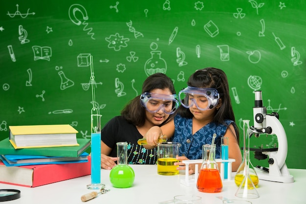 Duas lindas garotas indianas ou asiáticas experimentando ou estudando ciências em laboratório, sobre fundo de quadro verde com rabiscos educacionais