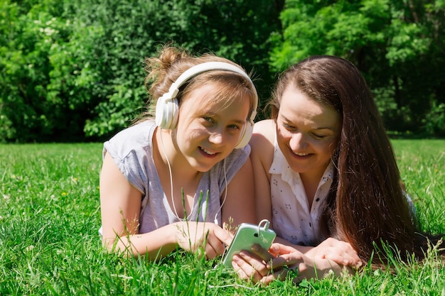 Duas lindas garotas mais jovens e mais velhas leem as notícias nas mídias sociais na grama do parque da cidade