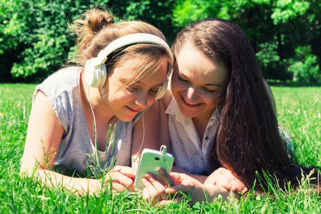 Duas lindas garotas mais jovens e mais velhas leem as notícias nas mídias sociais na grama do parque da cidade