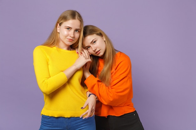 Duas lindas jovens loiras gêmeas meninas em roupas coloridas vivas em pé isoladas na parede de azul violeta pastel. Conceito de estilo de vida familiar de pessoas.